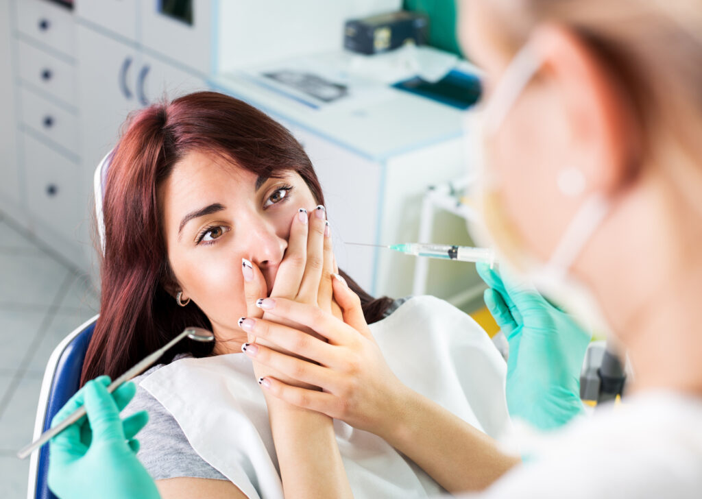 Young female dentist giving anesthesia to the patient before dental surgery. The patient in fear holds hands over mouth.